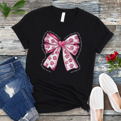 A black, coquette-style dog lover t-shirt with a bow design featuring a bow design filled with pink paw prints, surrounded by the script text 'dogs make everything better', displayed flat with a pair of blue denim shorts and white shoes on a wooden surface.
