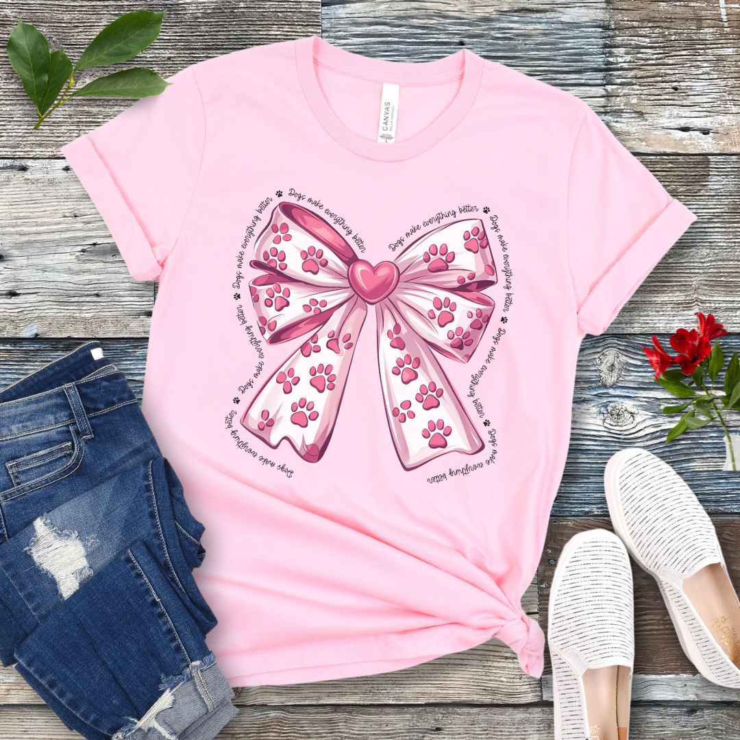 A pink, coquette-style dog lover t-shirt with a bow design featuring a bow design filled with pink paw prints, surrounded by the script text 'dogs make everything better', displayed flat with a pair of blue denim shorts and white shoes on a wooden surface.