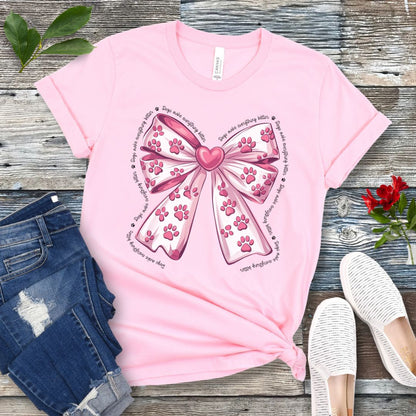 A pink, coquette-style dog lover t-shirt with a bow design featuring a bow design filled with pink paw prints, surrounded by the script text 'dogs make everything better', displayed flat with a pair of blue denim shorts and white shoes on a wooden surface.