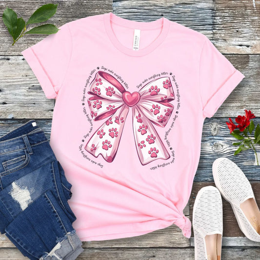 A pink, coquette-style dog lover t-shirt with a bow design featuring a bow design filled with pink paw prints, surrounded by the script text 'dogs make everything better', displayed flat with a pair of blue denim shorts and white shoes on a wooden surface.