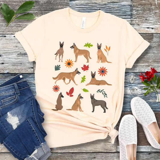 A beige Bella Canvas 3001 unisex short sleeve T-shirt with illustrations of Belgian Malinois dogs, fall leaves and flowers, laid out flat with jeans and casual shoes.