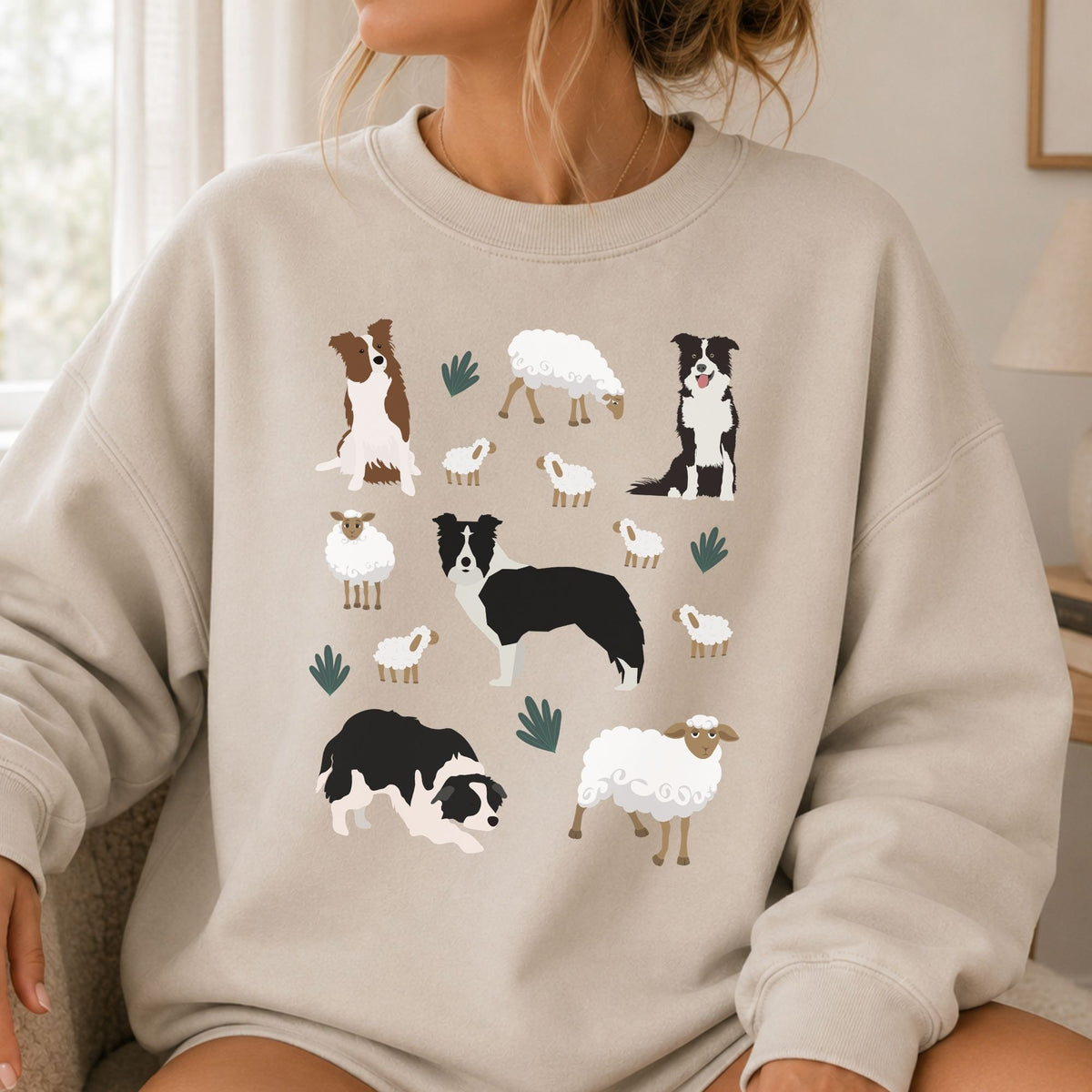 A woman wearing a light beige crewneck sweatshirt with a graphic print of various Border Collie dogs and sheep in black, white, and brown, set against a soft-focus indoor background.