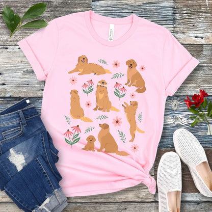 Pink dog lover t-shirt with cottagecore-style design made of Golden Retriever dogs in various positions, accented by flowers and leaves, shown on a wooden surface with jeans and shoes.