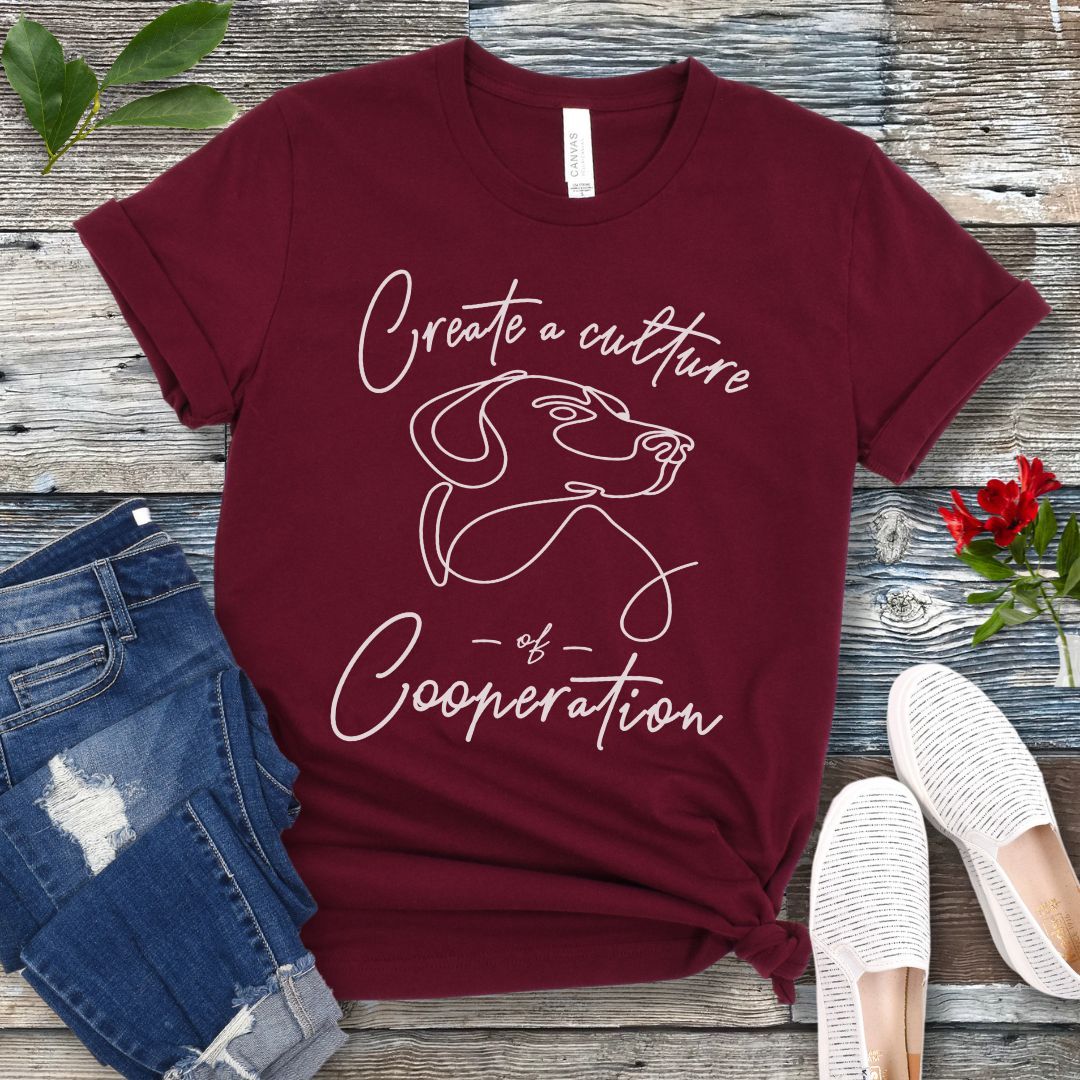 Maroon t-shirt with a dog illustration and positive dog training quote on a wooden surface with jeans and shoes.