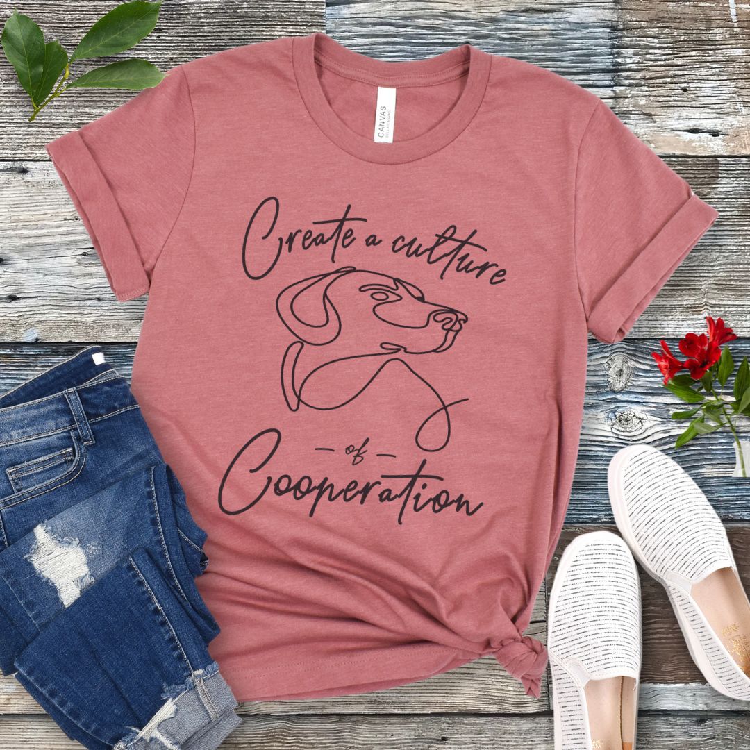 Mauve t-shirt with a dog illustration and positive dog training quote on a wooden surface with jeans and shoes.