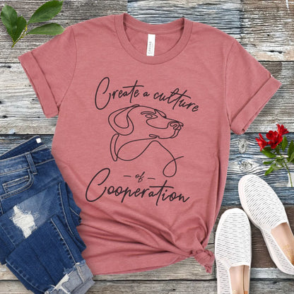 Mauve t-shirt with a dog illustration and positive dog training quote on a wooden surface with jeans and shoes.