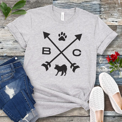 A grey t-shirt for Border Collie lovers with a graphic of crossed arrows, the letters BC, and the silhouette of a Border Collie dog in the center, displayed on a wooden surface with a pair of jeans and white shoes in the background. 