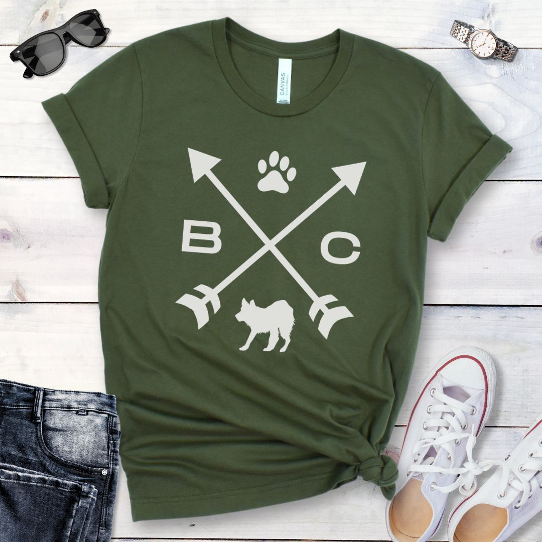 A military green t-shirt for Border Collie lovers with a graphic of crossed arrows, the letters BC, and the silhouette of a Border Collie dog in the center, displayed on a wooden surface with a pair of jeans and white shoes in the background. 