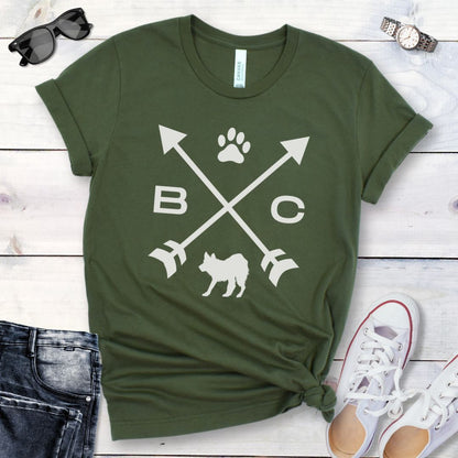 A military green t-shirt for Border Collie lovers with a graphic of crossed arrows, the letters BC, and the silhouette of a Border Collie dog in the center, displayed on a wooden surface with a pair of jeans and white shoes in the background. 