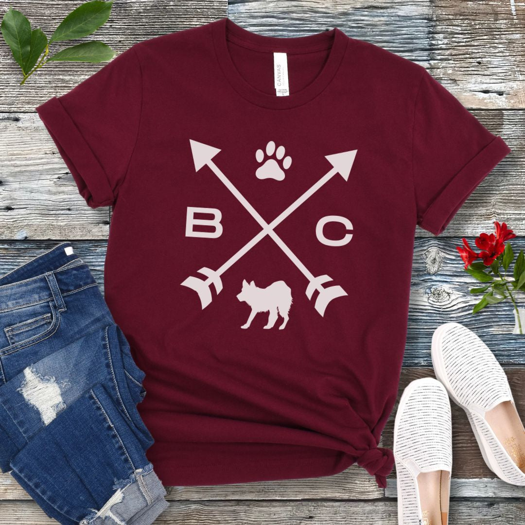 A maroon t-shirt for Border Collie lovers with a graphic of crossed arrows, the letters BC, and the silhouette of a Border Collie dog in the center, displayed on a wooden surface with a pair of jeans and white shoes in the background. 