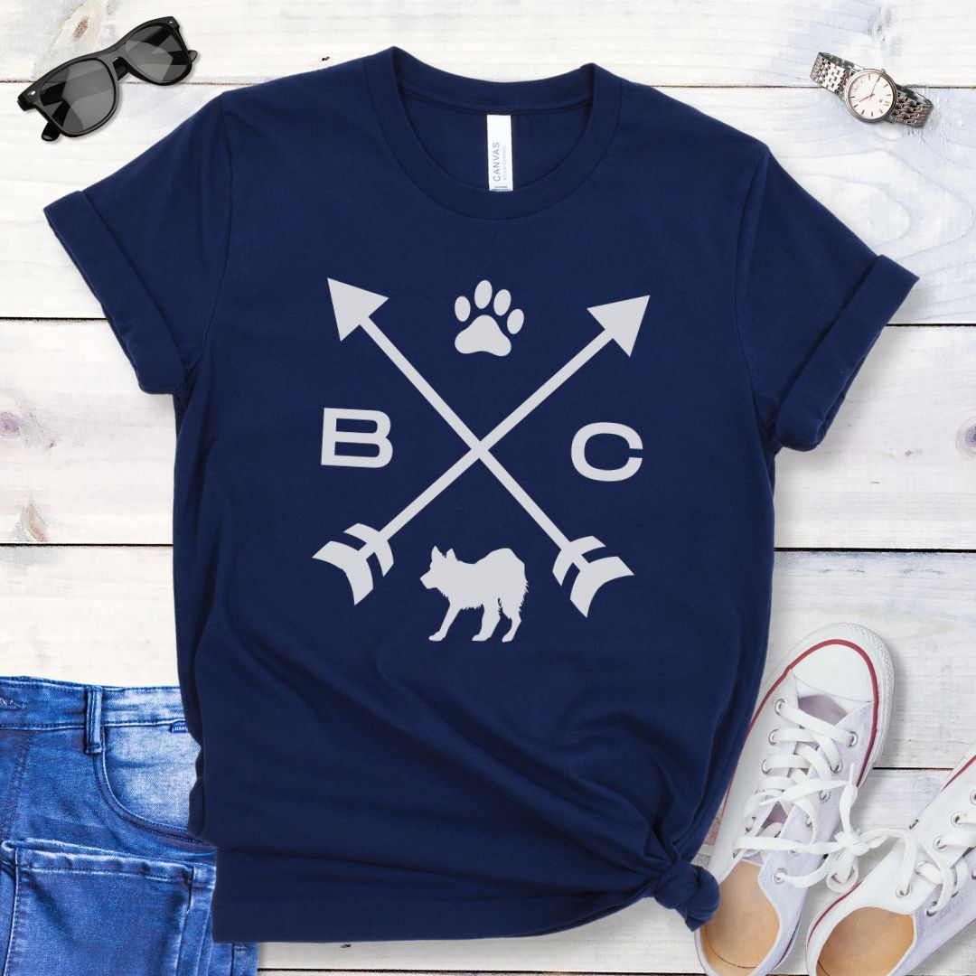 A navy blue t-shirt for Border Collie lovers with a graphic of crossed arrows, the letters BC, and the silhouette of a Border Collie dog in the center, displayed on a wooden surface with a pair of jeans and white shoes in the background. 