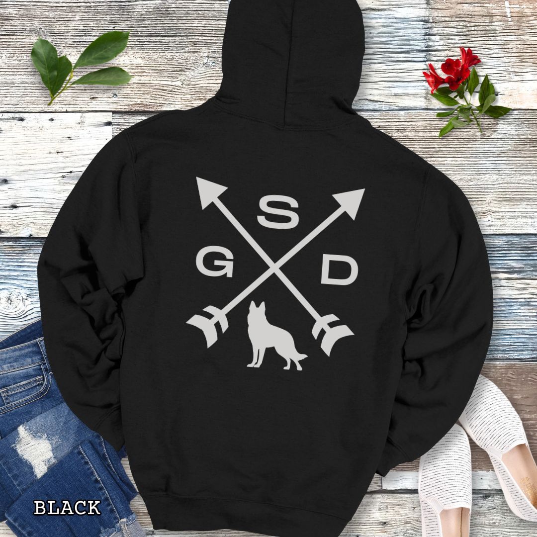 Back view of a black unisex hoodie with a crossed arrows design with 'GSD' text and a German Shepherd silhouette, presented on a wooden surface with accessories.