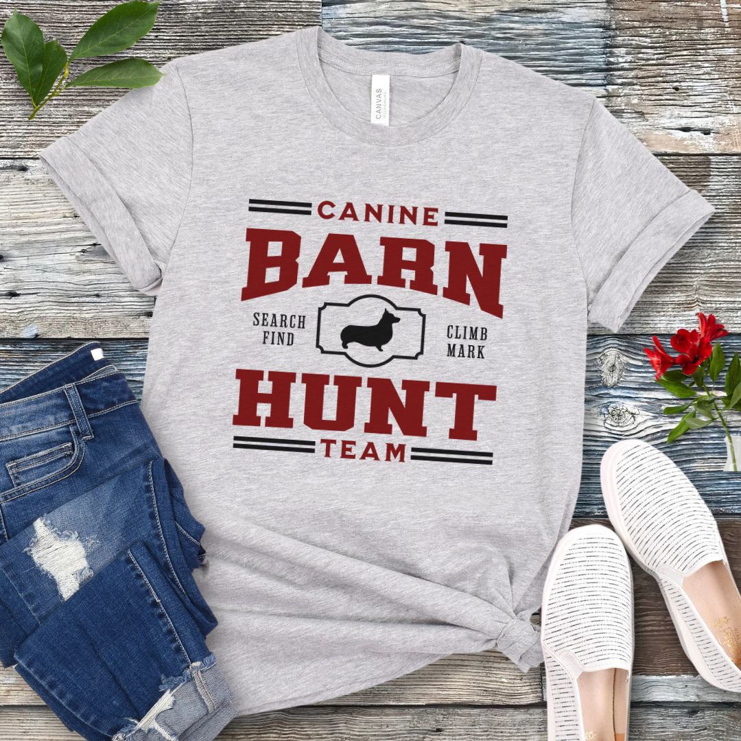 Gray dog sports t-shirt with red and black 'Canine Barn Hunt Team' text and custom dog breed silhouette on a wooden surface with jeans and shoes.