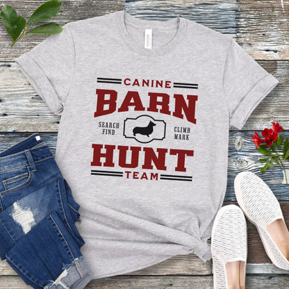Gray dog sports t-shirt with red and black 'Canine Barn Hunt Team' text and custom dog breed silhouette on a wooden surface with jeans and shoes.