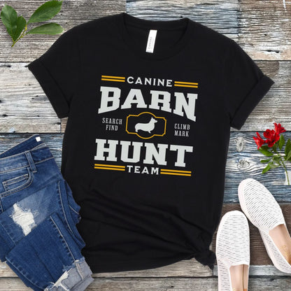 Black dog sports t-shirt with white and gold 'Canine Barn Hunt Team' text and custom dog breed silhouette on a wooden surface with jeans and shoes.