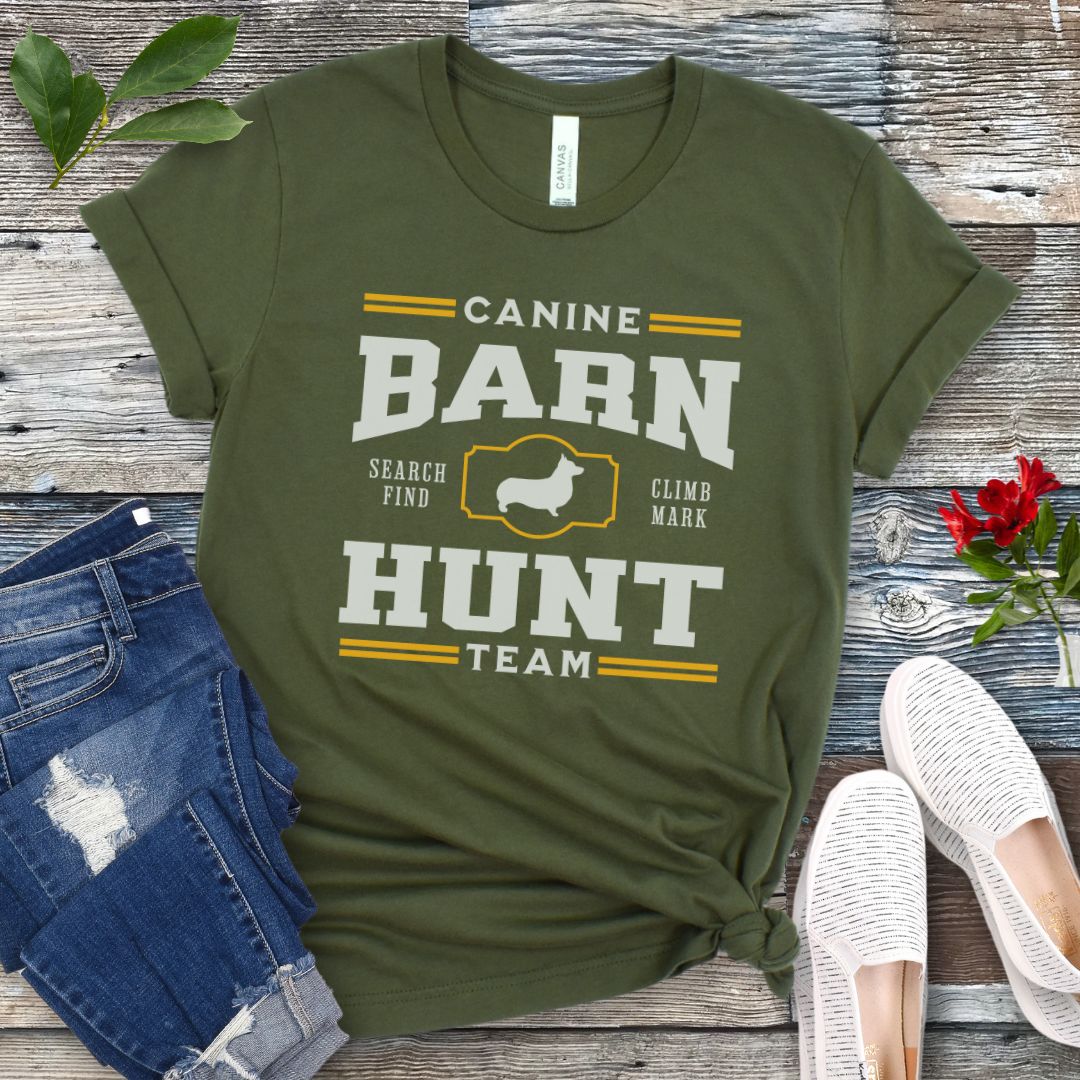 Military green dog sports t-shirt with gold and white 'Canine Barn Hunt Team' text and custom dog breed silhouette on a wooden surface with jeans and shoes.