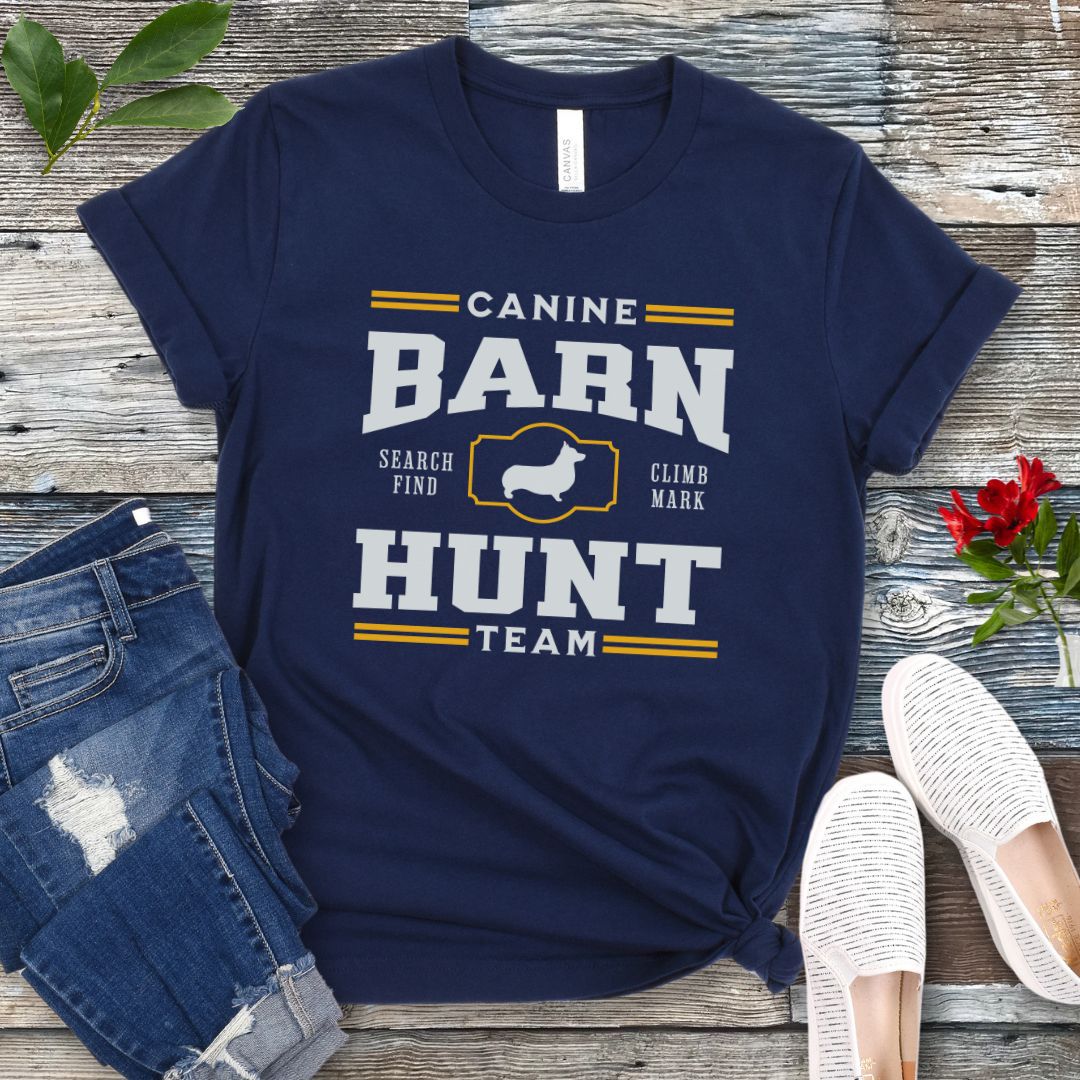 Navy blue dog sports t-shirt with gold and white 'Canine Barn Hunt Team' text and custom dog breed silhouette on a wooden surface with jeans and shoes.
