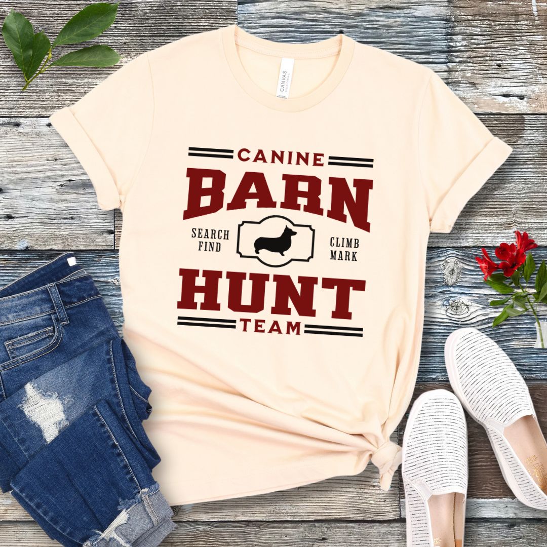 Beige dog sports t-shirt with red and black 'Canine Barn Hunt Team' text and custom dog breed silhouette on a wooden surface with jeans and shoes.