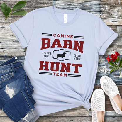 Light blue dog sports t-shirt with red and black 'Canine Barn Hunt Team' text and custom dog breed silhouette on a wooden surface with jeans and shoes.