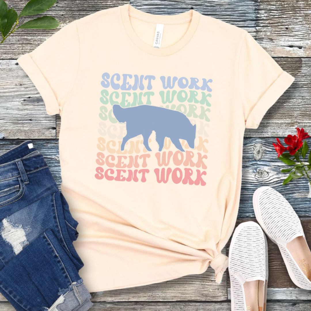 Beige dog scent training t-shirt with wavy multicolored 'SCENT WORK' text in a retro-style font and a custom dog breed silhouette, displayed flat with a pair of blue jeans and white shoes.