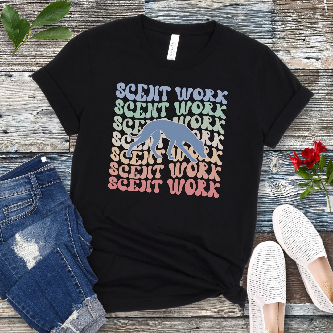 Black dog scent training t-shirt with wavy multicolored 'SCENT WORK' text in a retro-style font and a custom dog breed silhouette, displayed flat with a pair of blue jeans and white shoes.
