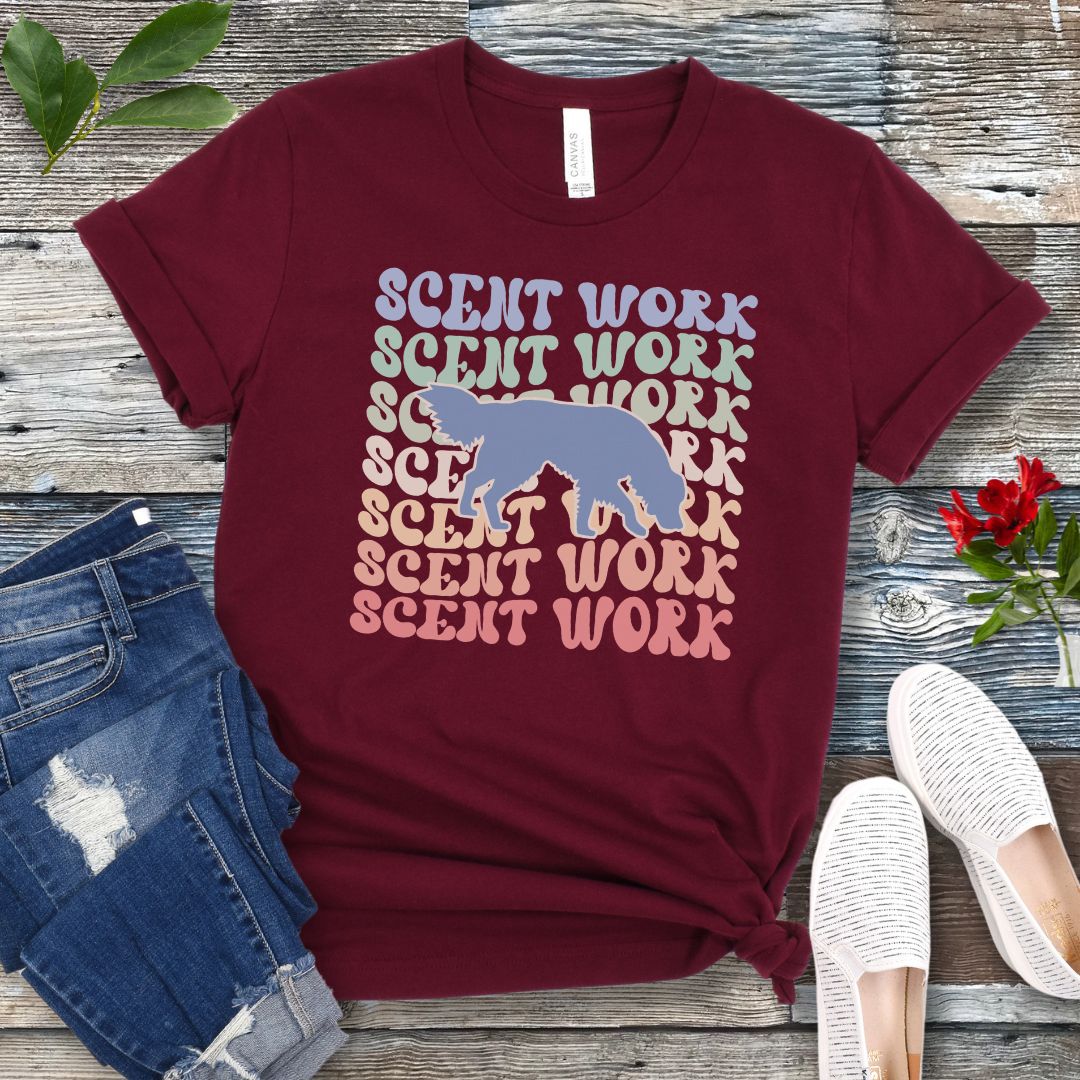 Maroon dog scent training t-shirt with wavy multicolored 'SCENT WORK' text in a retro-style font and a custom dog breed silhouette, displayed flat with a pair of blue jeans and white shoes.