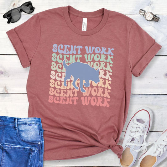 Mauve dog scent training t-shirt with wavy multicolored 'SCENT WORK' text in a retro-style font and a custom dog breed silhouette, displayed flat with a pair of blue jeans and white shoes.