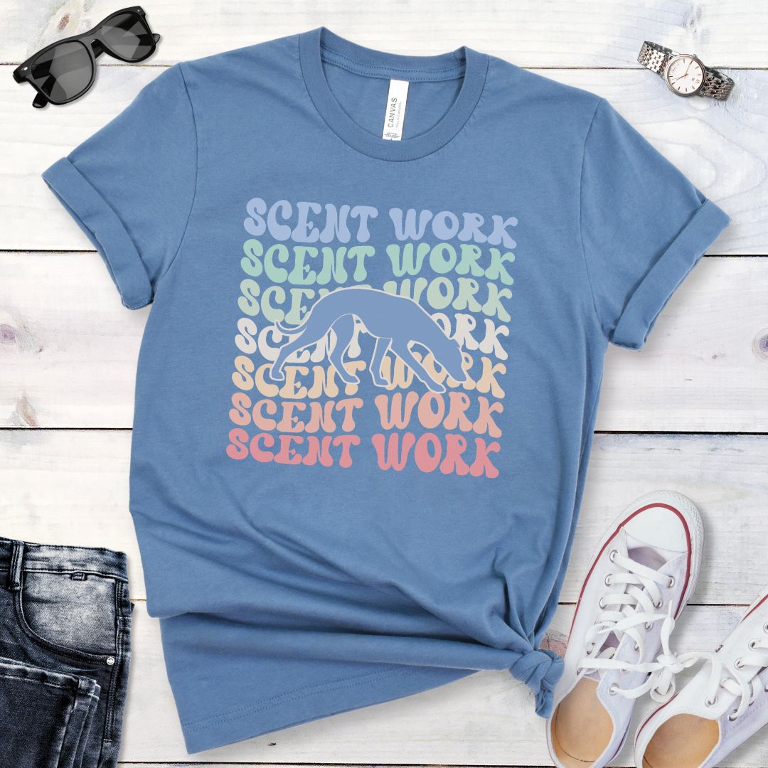 Steel blue dog scent training t-shirt with wavy multicolored 'SCENT WORK' text in a retro-style font and a custom dog breed silhouette, displayed flat with a pair of blue jeans and white shoes.