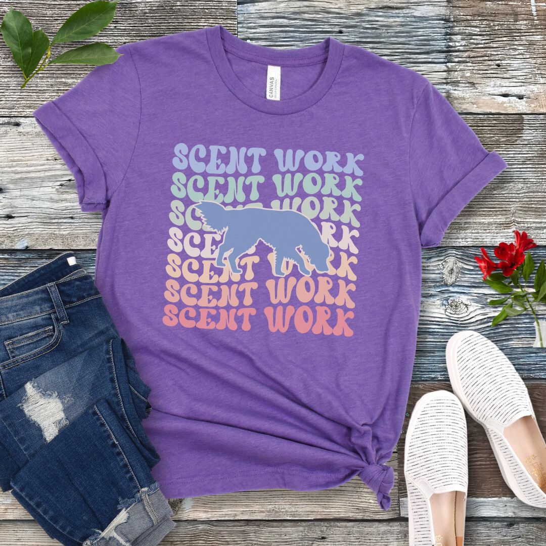 Purple dog scent training t-shirt with wavy multicolored 'SCENT WORK' text in a retro-style font and a custom dog breed silhouette, displayed flat with a pair of blue jeans and white shoes.