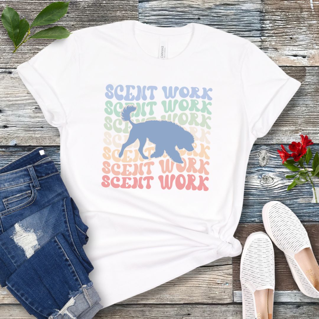 White dog scent training t-shirt with wavy multicolored 'SCENT WORK' text in a retro-style font and a custom dog breed silhouette, displayed flat with a pair of blue jeans and white shoes.