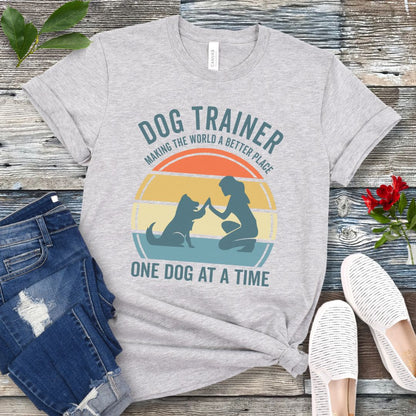 Light grey t-shirt with 'Dog Trainer' graphic and positive quote on a wooden surface with jeans and shoes.