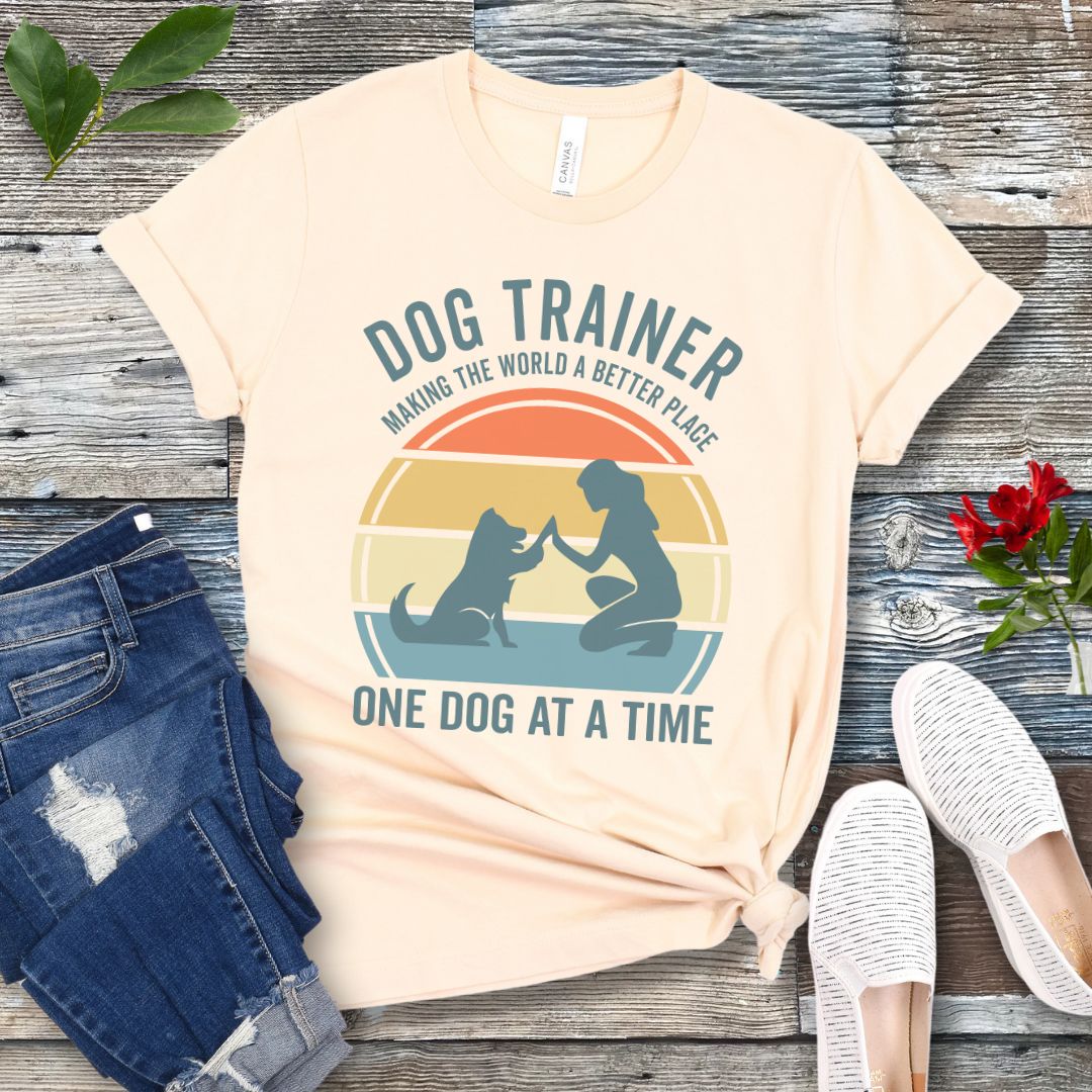 Light beige t-shirt with 'Dog Trainer' graphic and positive quote on a wooden surface with jeans and shoes.