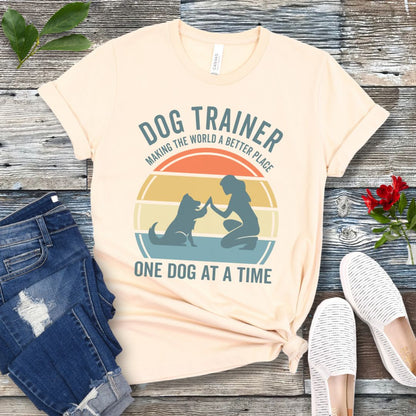 Light beige t-shirt with 'Dog Trainer' graphic and positive quote on a wooden surface with jeans and shoes.