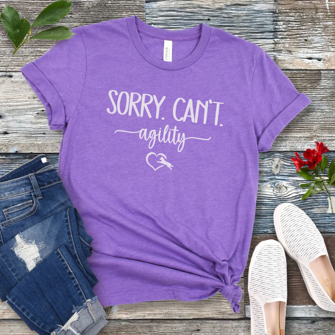 A purple dog agility t-shirt with the text 'SORRY, CAN'T. agility' text and a dog leaping into a heart graphic, displayed flat with a pair of blue denim jeans and white shoes.