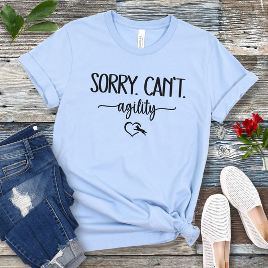 A light blue dog agility t-shirt with the text 'SORRY, CAN'T. agility' text and a dog leaping into a heart graphic, displayed flat with a pair of blue denim jeans and white shoes.