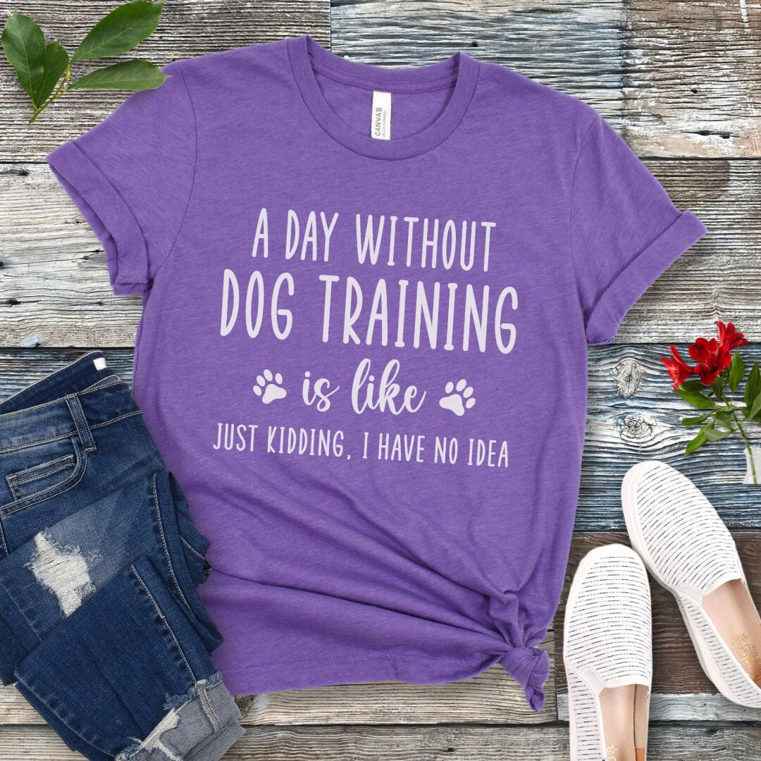 Purple unisex t-shirt with humorous dog training quote, 'A Day Without Dog Training Is Like, Just Kidding, I have No Idea' text, presented on a wooden surface with jeans and shoes.