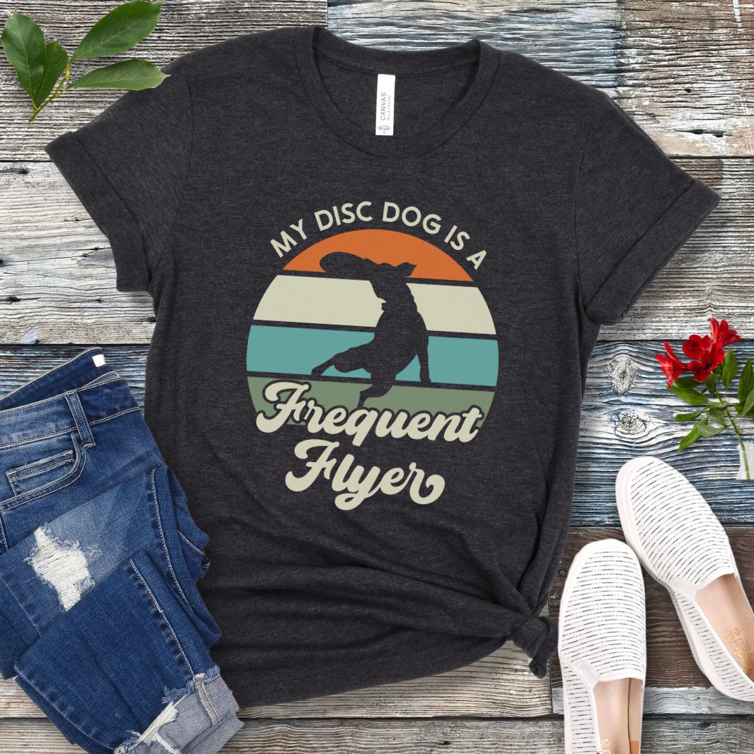 Dark grey unisex disc dog t-shirt with colorful vintage sunset, leaping dog catching a disc, and the humorous text 'My Disc dog is a frequent flyer'. 