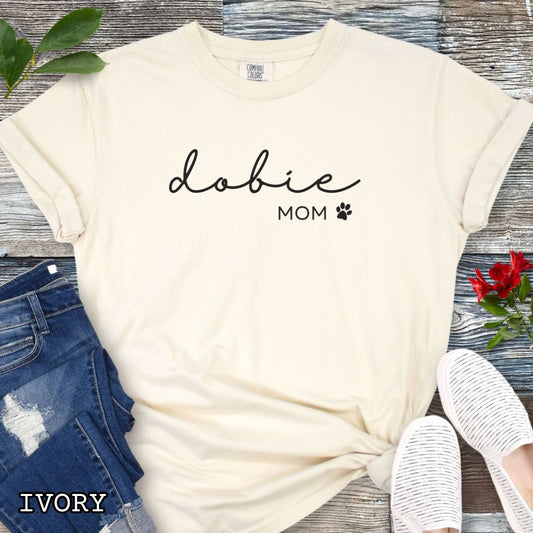 Beige Comfort Colors t-shirt for Doberman dog lovers with 'dobie mom' text design in feminine handwritten script, accompanied by a small paw print, shown on a wooden surface with sunglasses, jeans, and sneakers.