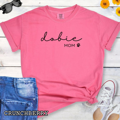 Pink Comfort Colors t-shirt for Doberman dog lovers with 'dobie mom' text design in feminine handwritten script, accompanied by a small paw print, shown on a wooden surface with sunglasses, jeans, and sneakers.