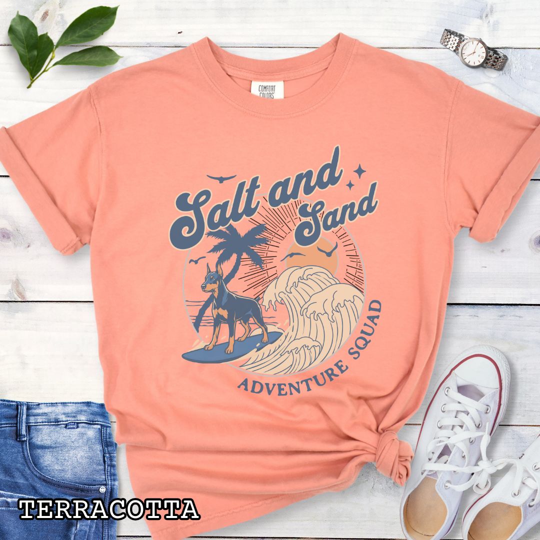 Orange Comfort Colors t-shirt for Doberman Pinscher dog lovers with retro surfing Doberman dog graphic in a beach setting with 'Salt and Sand Adventure Squad' text, shown on a wooden surface.