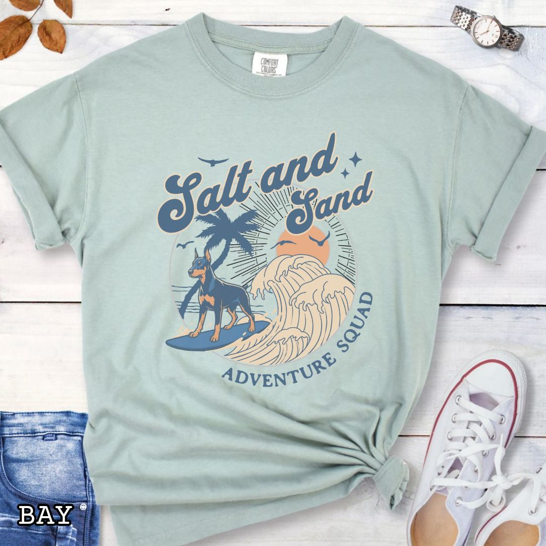Pale green Doberman Pinscher dog lovers with retro surfing Doberman dog graphic in a beach setting with 'Salt and Sand Adventure Squad' text, shown on a wooden surface.