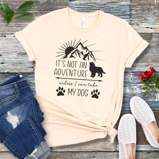 A beige dog lover T-shirt with a graphic that says 'IT'S NOT AN ADVENTURE UNLESS I CAN TAKE MY DOG' accompanied by images of a mountain, a dog and paw prints. The shirt is displayed alongside a pair of jeans and a pair of white shoes on a wooden background.