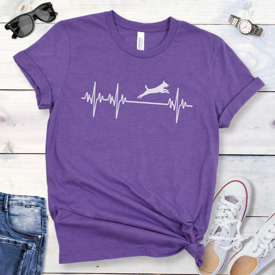 Purple dog agility t-shirt with a heartbeat/EKG design with leaping dog on a wooden surface with jeans and shoes.