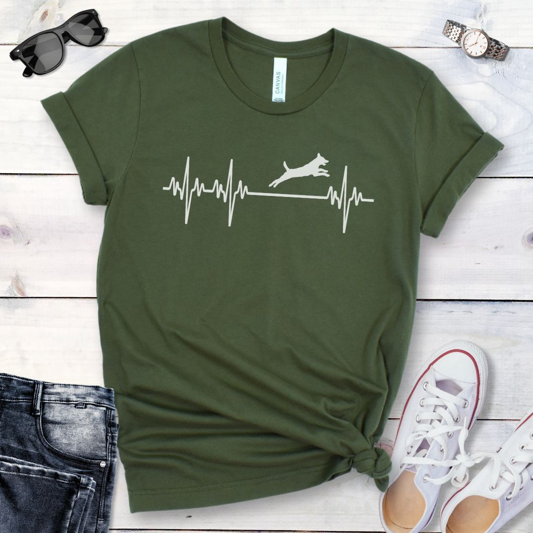Military green dog agility t-shirt with a heartbeat/EKG design with leaping dog on a wooden surface with jeans and shoes.