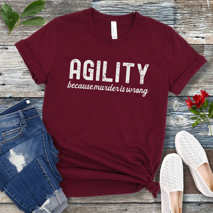 Maroon t-shirt with 'AGILITY because murder is wrong' text on a wooden surface with jeans and shoes.