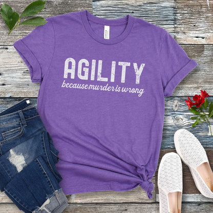 Purple t-shirt with 'AGILITY because murder is wrong' text on a wooden surface with jeans and shoes.