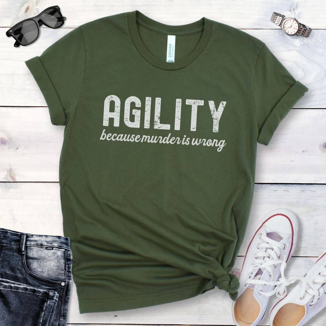 Military green dog agility t-shirt with 'AGILITY because murder is wrong' text on a wooden surface with jeans and shoes.