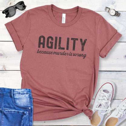 Mauve dog agility t-shirt with 'AGILITY because murder is wrong' text on a wooden surface with jeans and shoes.