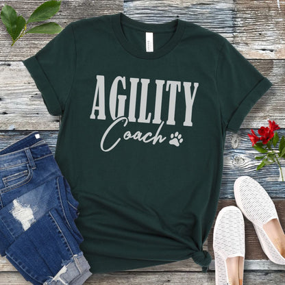 Dark green dog agility training t-shirt with 'AGILITY Coach' text in bold modern lettering, with accompanying paw print, shown on a wooden surface with jeans and shoes.