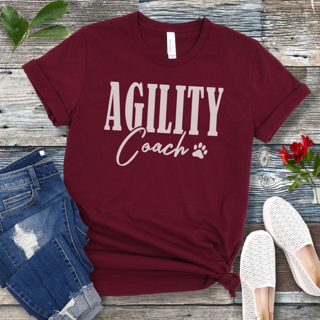 Maroon dog agility training t-shirt with 'AGILITY Coach' text in bold modern lettering, with accompanying paw print, shown on a wooden surface with jeans and shoes.